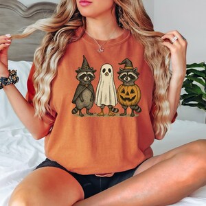 May include: Orange t-shirt featuring a Halloween-themed graphic. The graphic depicts three cartoon characters: a raccoon in a witch hat and cape, a ghost, and a raccoon in a pumpkin costume. The shirt is a solid orange color.