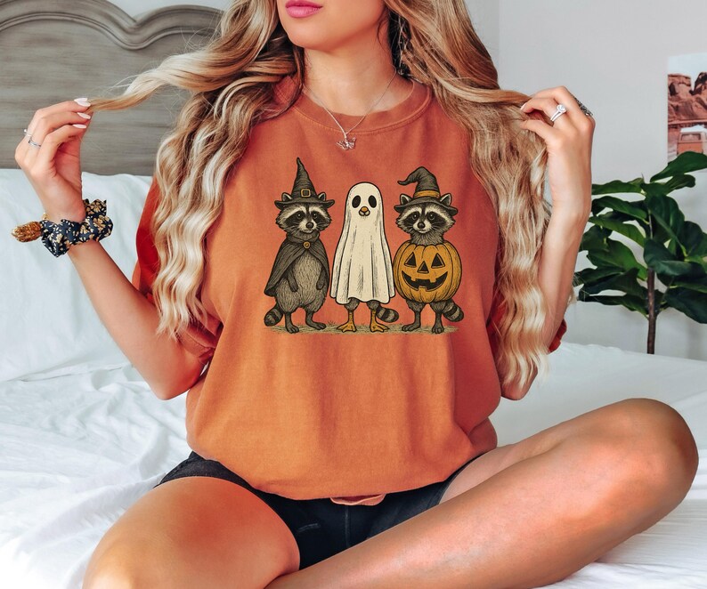 May include: Orange t-shirt featuring a Halloween-themed graphic. The graphic depicts three cartoon characters: a raccoon in a witch hat and cape, a ghost, and a raccoon in a pumpkin costume. The shirt is a solid orange color.