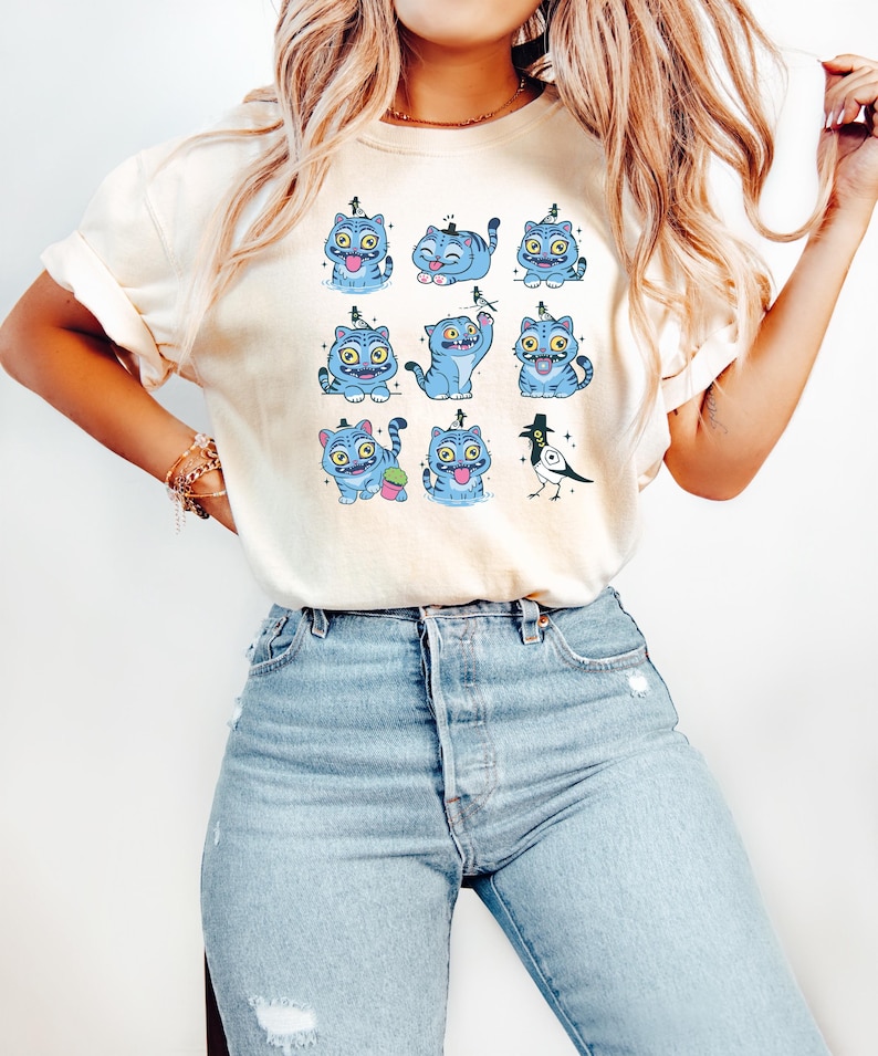May include: Cream-colored t-shirt featuring a grid of cartoon blue cats with various expressions and poses. The cats have yellow eyes and are decorated with small hats. A black and white bird with a top hat is also featured. The shirt is paired with blue jeans.
