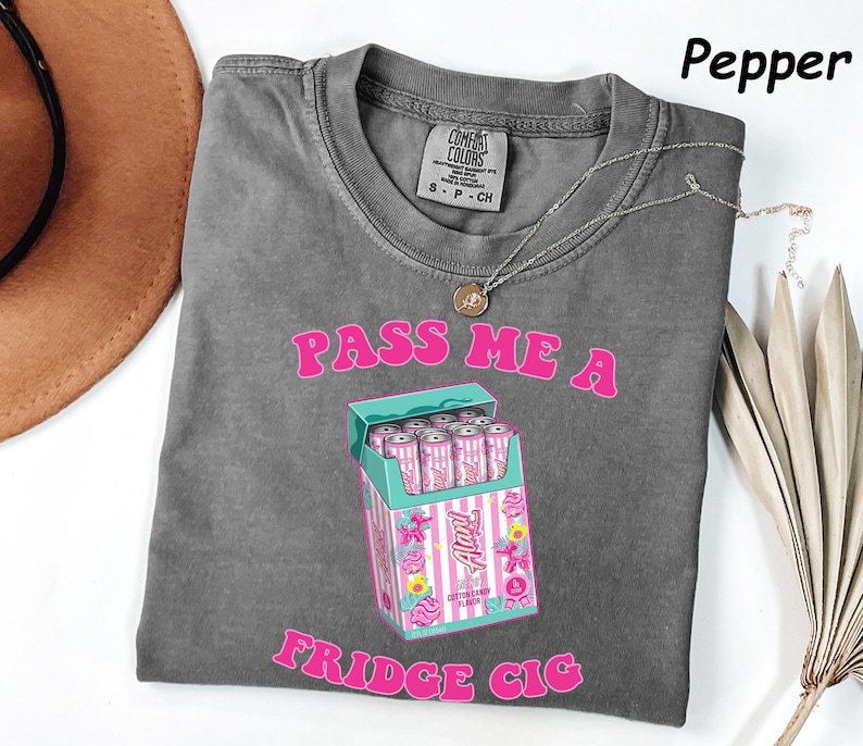 May include: A gray Comfort Colors t-shirt with the text "PASS ME A FRIDGE CIG" in pink. The graphic is a carton of pink and white cans. A gold necklace is draped on the shirt.