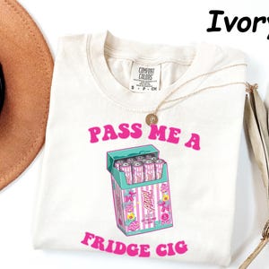 May include: Ivory-colored t-shirt with the text "PASS ME A FRIDGE CIG" in pink. The graphic features a teal cigarette box with pink and white striped cigarettes. A gold necklace and a brown hat are also visible.