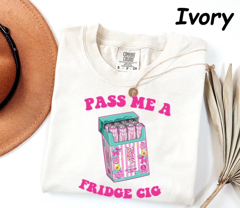May include: Ivory-colored t-shirt with the text "PASS ME A FRIDGE CIG" in pink. The graphic features a teal cigarette box with pink and white striped cigarettes. A gold necklace and a brown hat are also visible.
