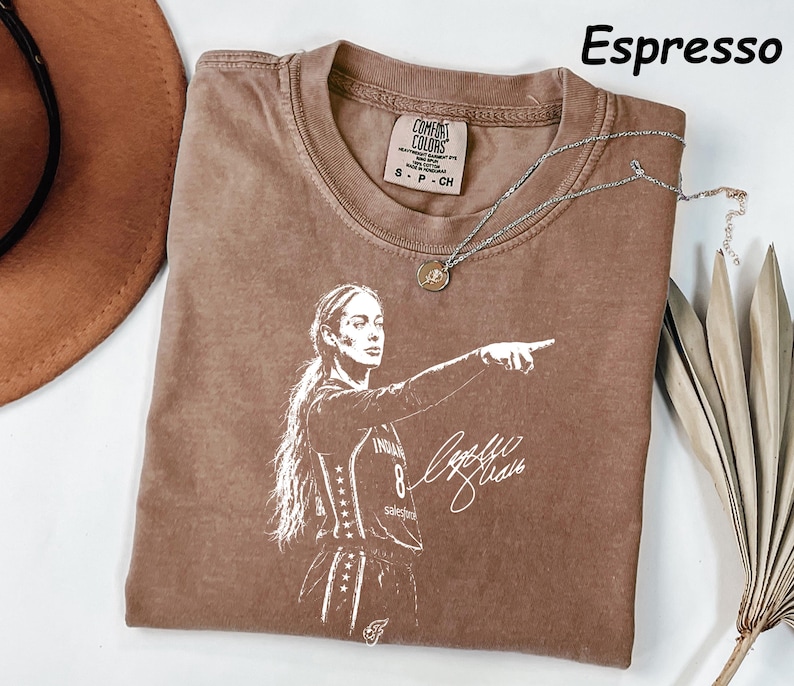 May include: An espresso-colored t-shirt featuring a white graphic print of a basketball player pointing with a signature below. The shirt has a crew neckline and a Comfort Colors brand label at the inner neck. The text Espresso is visible.