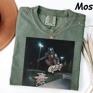 May include: Moss green t-shirt featuring a graphic of The Mandalorian and Grogu skateboarding at night. The shirt has a crew neck and a small tag on the upper back. A gold necklace is draped across the shirt. The word "Moss" is in the upper right corner.
