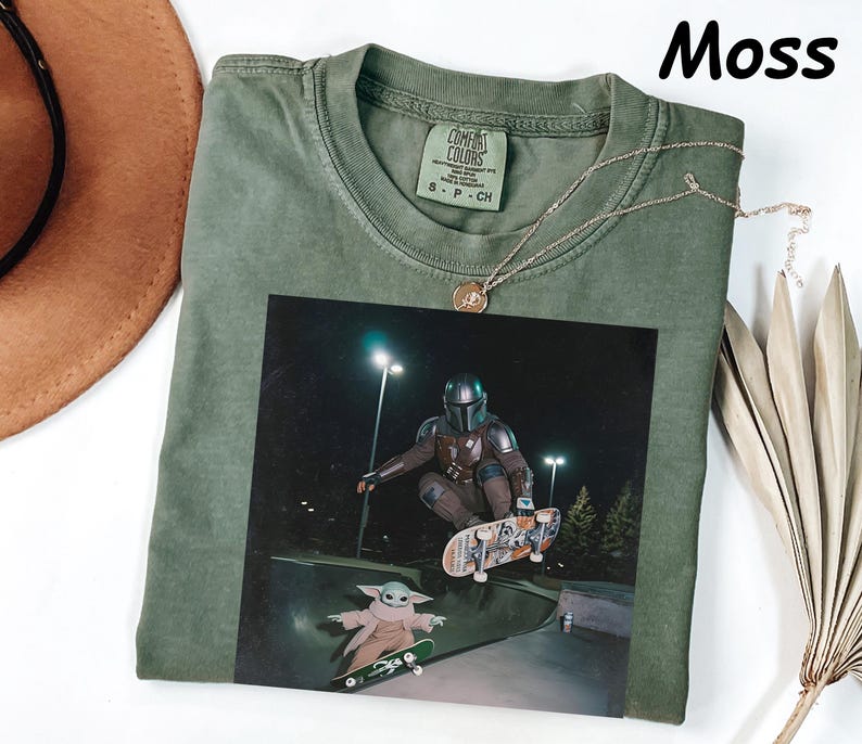 May include: Moss green t-shirt featuring a graphic of The Mandalorian and Grogu skateboarding at night. The shirt has a crew neck and a small tag on the upper back. A gold necklace is draped across the shirt. The word "Moss" is in the upper right corner.