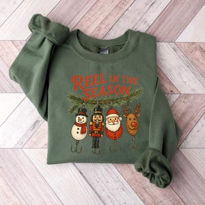 May include: Green sweatshirt with the text "REEL IN THE SEASON" in red. The graphic features Christmas-themed fishing lures: a snowman, nutcracker, Santa, and reindeer. The sweatshirt is displayed on a light wood-grain surface.