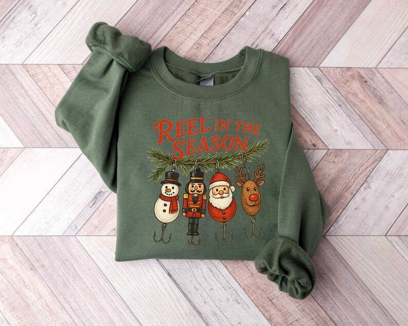 May include: Green sweatshirt with the text "REEL IN THE SEASON" in red. The graphic features Christmas-themed fishing lures: a snowman, nutcracker, Santa, and reindeer. The sweatshirt is displayed on a light wood-grain surface.