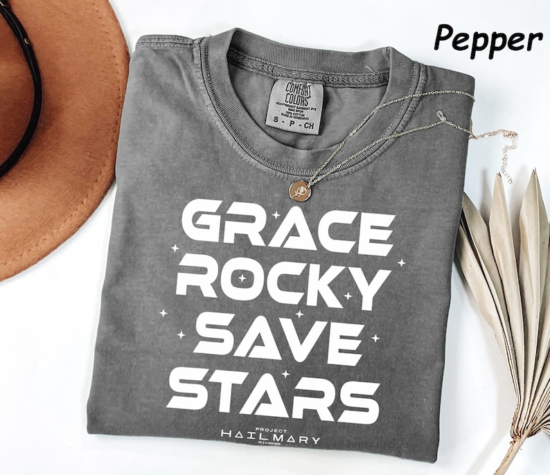 May include: A gray t-shirt with the words "GRACE ROCKY SAVE STARS" in white block letters. The shirt has a crew neck and short sleeves. A gold necklace with a pendant rests on the shirt. The word "Pepper" is in the upper right corner.