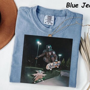 May include: Pale blue t-shirt with a graphic of a character from The Mandalorian skateboarding. The image includes the text "Blue Jean" at the top. A gold necklace is draped across the shirt. The shirt is labeled "Comfort Colors" on the tag.