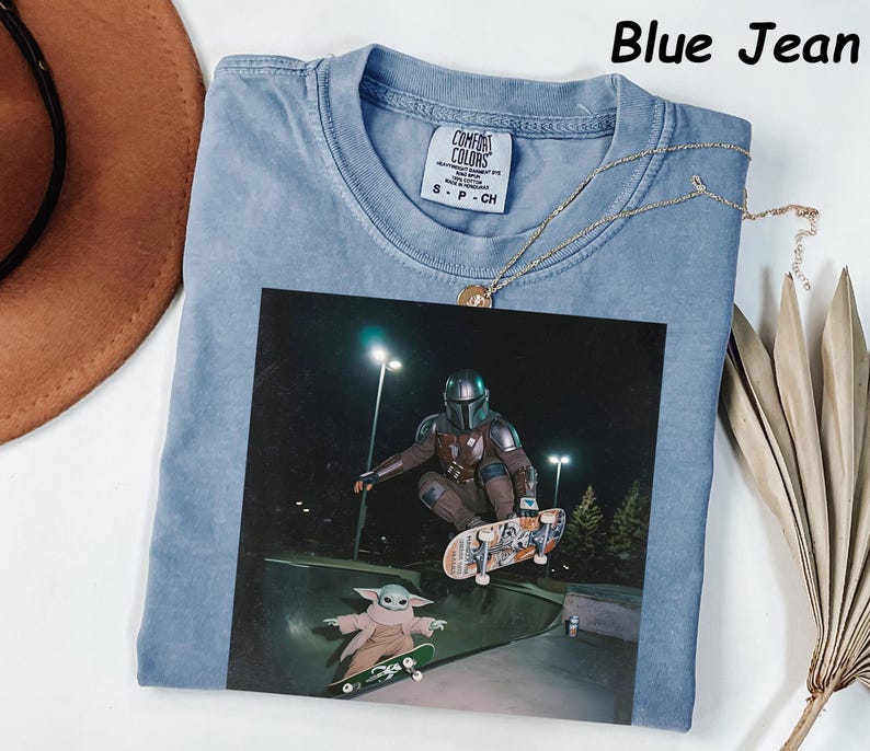 May include: Pale blue t-shirt with a graphic of a character from The Mandalorian skateboarding. The image includes the text "Blue Jean" at the top. A gold necklace is draped across the shirt. The shirt is labeled "Comfort Colors" on the tag.