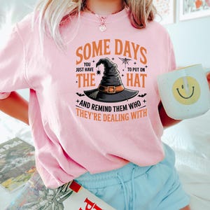 Puede incluir: Una camiseta rosa claro con el texto "SOME DAYS YOU JUST HAVE TO PUT ON THE HAT AND REMIND THEM WHO THEY'RE DEALING WITH." El gr&aacute;fico presenta un sombrero de bruja, una ara&ntilde;a y murci&eacute;lagos. La persona sostiene una taza blanca con una carita sonriente amarilla.