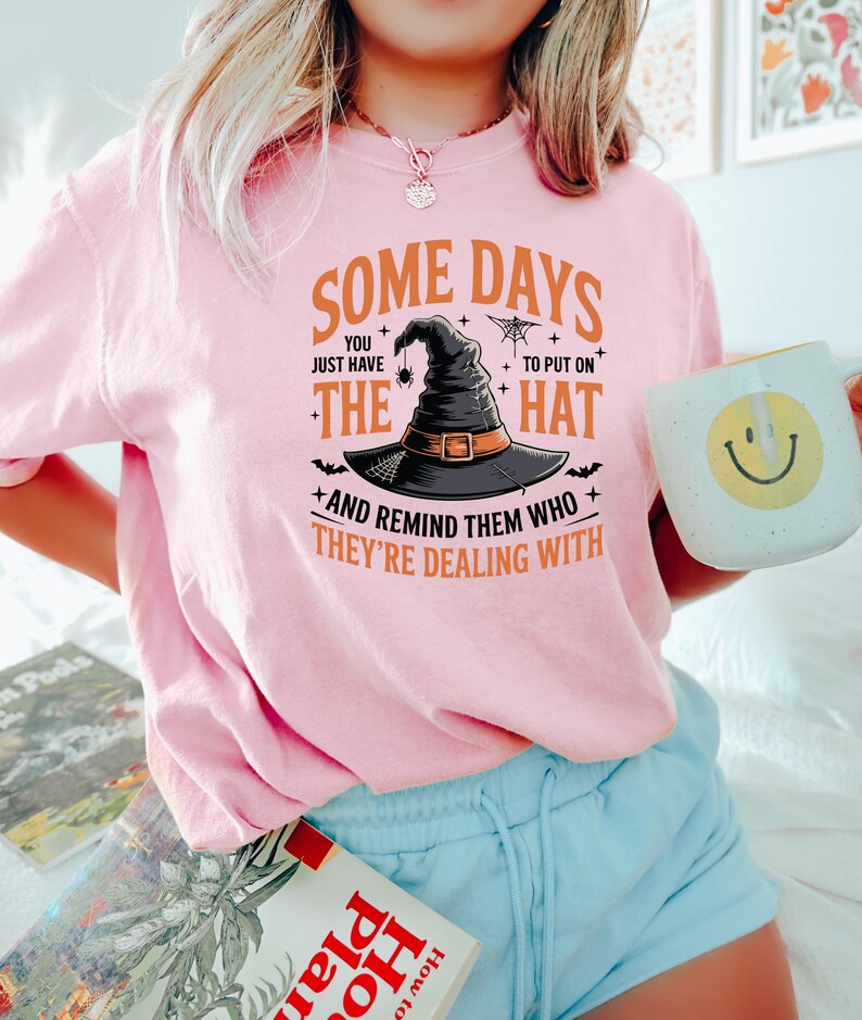 Puede incluir: Una camiseta rosa claro con el texto "SOME DAYS YOU JUST HAVE TO PUT ON THE HAT AND REMIND THEM WHO THEY'RE DEALING WITH." El gr&aacute;fico presenta un sombrero de bruja, una ara&ntilde;a y murci&eacute;lagos. La persona sostiene una taza blanca con una carita sonriente amarilla.