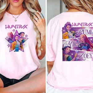 May include: Light pink t-shirt featuring a graphic of a group of animated characters inside a star, with the word "HUNTRAX" above. The back of the shirt displays the names RUMI, MIRA, and ZOEY with character illustrations.