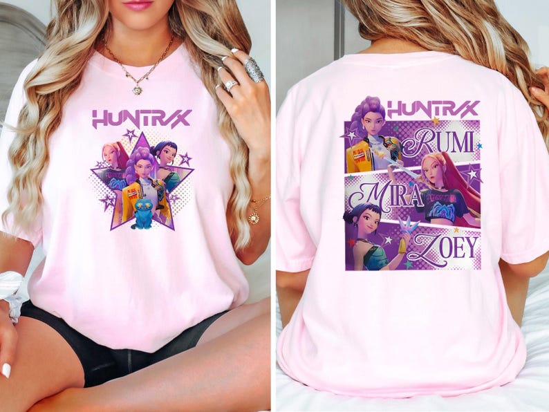 May include: Light pink t-shirt featuring a graphic of a group of animated characters inside a star, with the word "HUNTRAX" above. The back of the shirt displays the names RUMI, MIRA, and ZOEY with character illustrations.