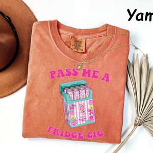May include: A coral-colored t-shirt with the text "PASS ME A FRIDGE CIG" in pink. The graphic features a cartoon cigarette pack with pink cans. A gold necklace and a brown hat are also visible.