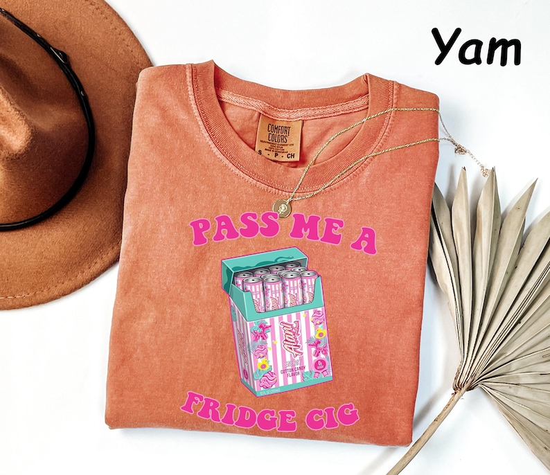 May include: A coral-colored t-shirt with the text "PASS ME A FRIDGE CIG" in pink. The graphic features a cartoon cigarette pack with pink cans. A gold necklace and a brown hat are also visible.