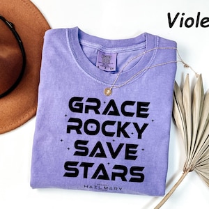 May include: A violet Comfort Colors t-shirt with the words "GRACE ROCKY SAVE STARS" in black. A gold necklace and a brown hat are next to the shirt. The text "Violet" is in the upper right corner.