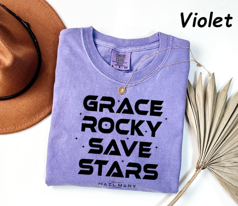 May include: A violet Comfort Colors t-shirt with the words "GRACE ROCKY SAVE STARS" in black. A gold necklace and a brown hat are next to the shirt. The text "Violet" is in the upper right corner.
