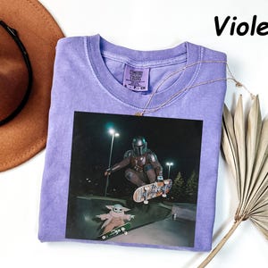 May include: A violet-coloured t-shirt with a graphic of The Mandalorian and Grogu skateboarding. The image is printed on a Comfort Colors shirt. A gold necklace is draped across the shirt. The word "Violet" is printed in black in the upper right corner.