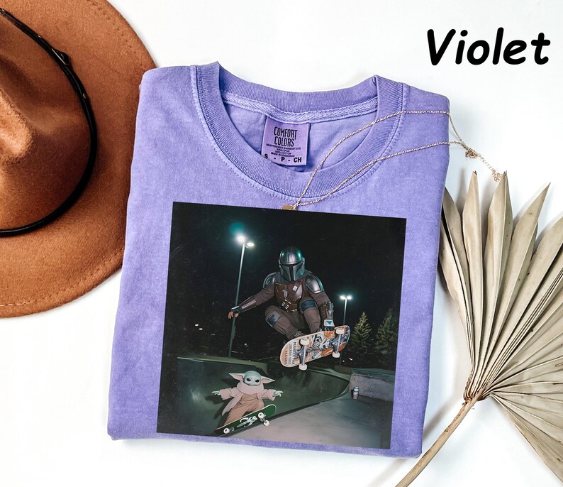 May include: A violet-coloured t-shirt with a graphic of The Mandalorian and Grogu skateboarding. The image is printed on a Comfort Colors shirt. A gold necklace is draped across the shirt. The word "Violet" is printed in black in the upper right corner.
