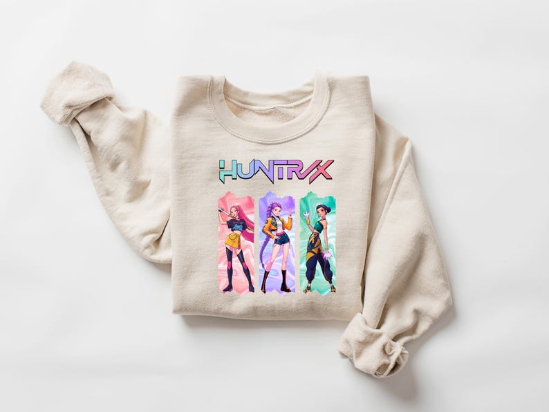 May include: Beige sweatshirt with the word "HUNTRX" in a colorful, gradient font. Below the text are three anime-style figures in colorful outfits. The sweatshirt is folded with the sleeves rolled up.