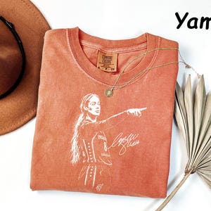 May include: A burnt orange crew neck t-shirt featuring a white graphic print of a basketball player pointing, with the text "Yam" visible in the upper right corner. The shirt has a Comfort Colors brand label at the inner neckline with size S.