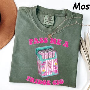 May include: Moss green t-shirt with the text "PASS ME A FRIDGE CIG" in pink. The graphic features a box of pink and white "Auntie" cotton candy flavored cans. A gold necklace is draped across the shirt.