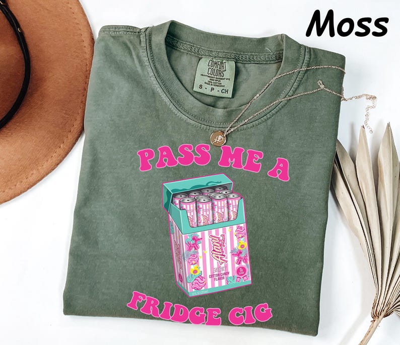 May include: Moss green t-shirt with the text "PASS ME A FRIDGE CIG" in pink. The graphic features a box of pink and white "Auntie" cotton candy flavored cans. A gold necklace is draped across the shirt.