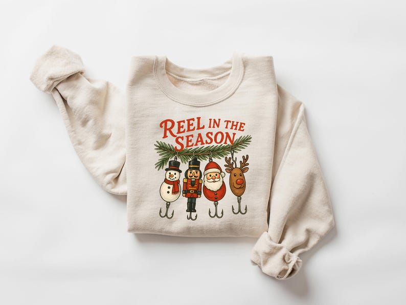 May include: Cream-colored sweatshirt with the text "REEL IN THE SEASON" in red. The design features four Christmas-themed fishing lures: a snowman, a nutcracker, Santa Claus, and a reindeer, all hanging from a green pine branch.