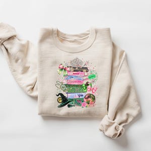 May include: Beige sweatshirt featuring a colorful graphic of stacked books, a witch's hat, and a crystal ball. The books have titles like "Popular" and "The Enchanted Forest." A tiara and pink bows add decorative elements to the design.