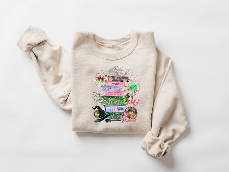 May include: Beige sweatshirt featuring a colorful graphic of stacked books, a witch's hat, and a crystal ball. The books have titles like "Popular" and "The Enchanted Forest." A tiara and pink bows add decorative elements to the design.