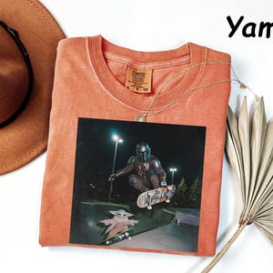 May include: A coral-coloured t-shirt featuring a graphic of a character from a popular science fiction series skateboarding. The image includes the text "Yam" in the upper right corner. A gold necklace and a brown hat are also visible.