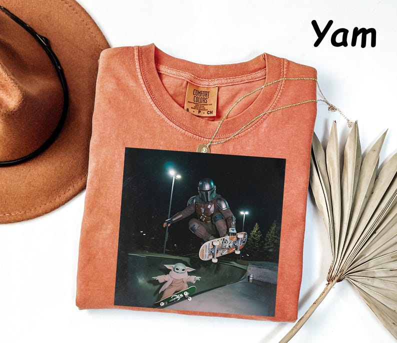 May include: A coral-coloured t-shirt featuring a graphic of a character from a popular science fiction series skateboarding. The image includes the text "Yam" in the upper right corner. A gold necklace and a brown hat are also visible.