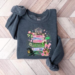 May include: Dark gray sweatshirt featuring a whimsical design of stacked books, a crown, and a witch's hat. The books have titles like "Popular" and "Driving Lessons". The design includes pink and green bows, a crystal ball, and stars.