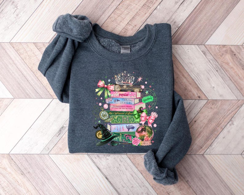 May include: Dark gray sweatshirt featuring a whimsical design of stacked books, a crown, and a witch's hat. The books have titles like "Popular" and "Driving Lessons". The design includes pink and green bows, a crystal ball, and stars.