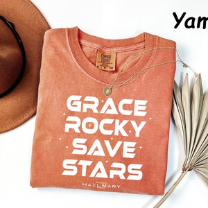 May include: A coral-colored t-shirt with the words "GRACE ROCKY SAVE STARS" in white block letters. A gold necklace and a brown hat are next to the shirt. The shirt has a crew neck and short sleeves. The text "PROJECT HAIL MARY" is at the bottom.