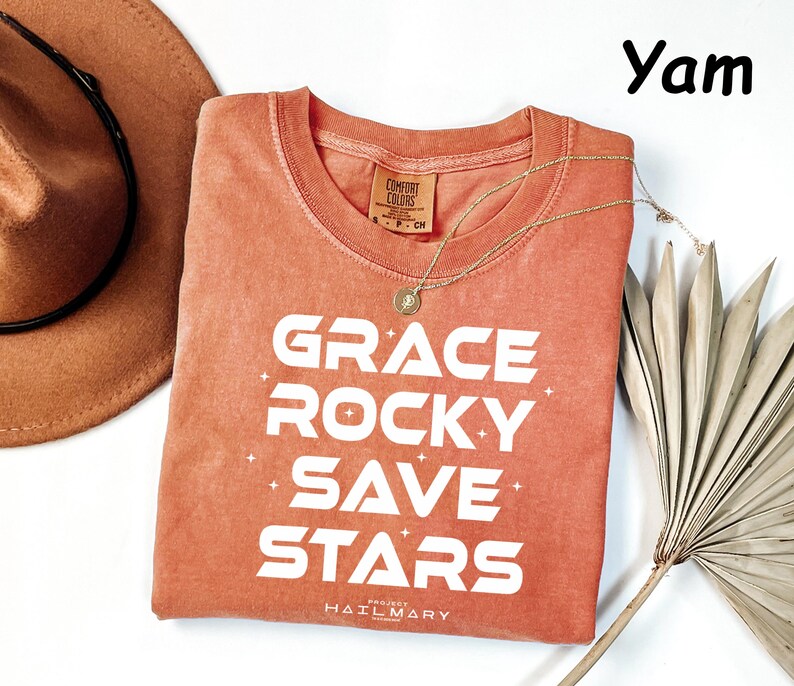 May include: A coral-colored t-shirt with the words "GRACE ROCKY SAVE STARS" in white block letters. A gold necklace and a brown hat are next to the shirt. The shirt has a crew neck and short sleeves. The text "PROJECT HAIL MARY" is at the bottom.