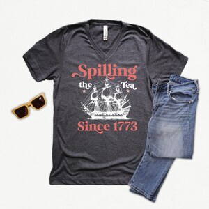 May include: Dark gray V-neck t-shirt with the text "Spilling the Tea Since 1773" and a white ship graphic. The image also includes blue jeans and wooden sunglasses.