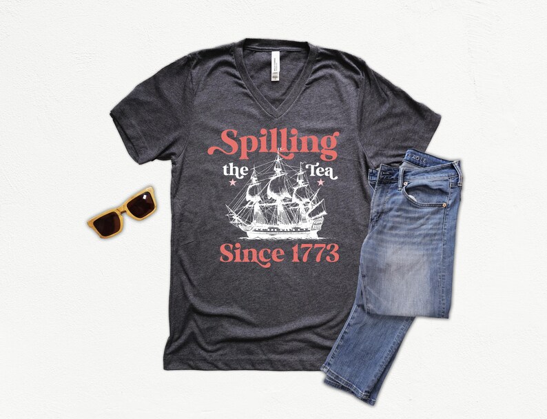 May include: Dark gray V-neck t-shirt with the text "Spilling the Tea Since 1773" and a white ship graphic. The image also includes blue jeans and wooden sunglasses.