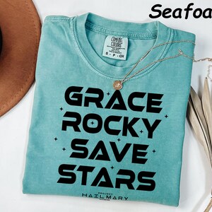 May include: Seafoam green t-shirt with the words "GRACE ROCKY SAVE STARS" in bold black letters. A gold necklace with a pendant is draped on the shirt. The shirt is folded on a white surface.