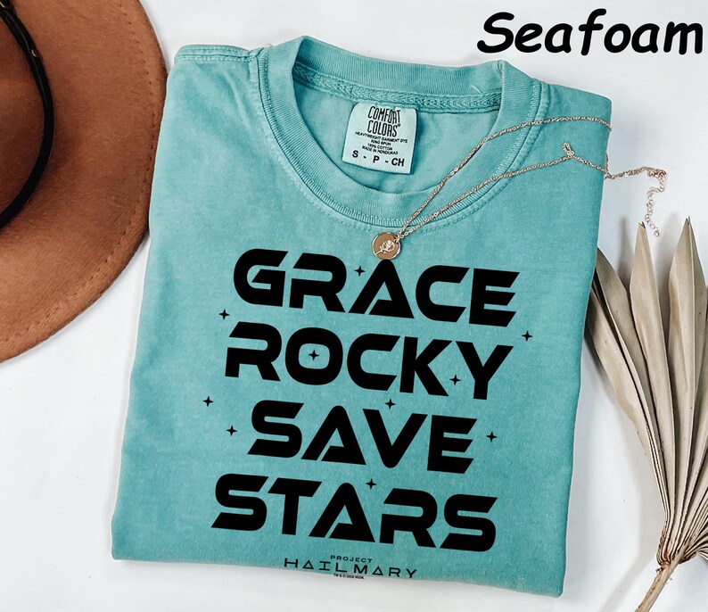 May include: Seafoam green t-shirt with the words "GRACE ROCKY SAVE STARS" in bold black letters. A gold necklace with a pendant is draped on the shirt. The shirt is folded on a white surface.