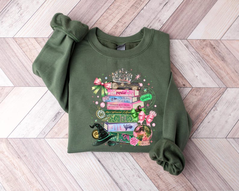 May include: Olive green sweatshirt featuring a stack of books with a crown, witch hat, and pink bows. The books have titles like "Popular" and "Wicked." The design includes a pink and green globe and a gold pocket watch. The sweatshirt is on a wooden background.