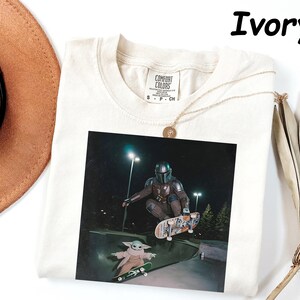 May include: Ivory-coloured t-shirt featuring a graphic of a character from the Mandalorian series skateboarding. The image shows the character in a skate park with another character. The shirt has a Comfort Colors tag.