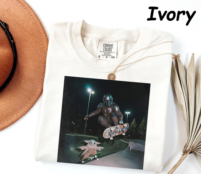 May include: Ivory-coloured t-shirt featuring a graphic of a character from the Mandalorian series skateboarding. The image shows the character in a skate park with another character. The shirt has a Comfort Colors tag.