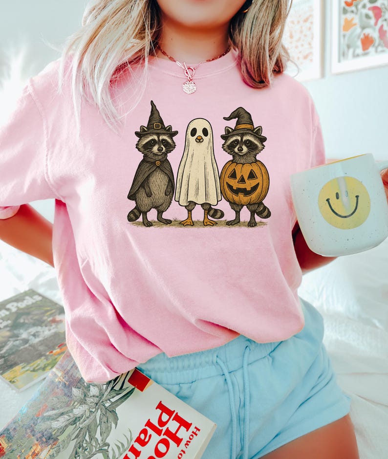May include: A light pink sweatshirt featuring a Halloween-themed graphic. The graphic depicts three cartoon characters: two raccoons wearing witch hats and a ghost. One raccoon has a pumpkin body. The sweatshirt is a comfortable, casual style.