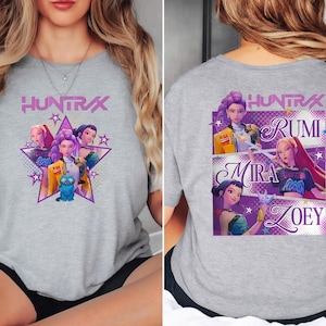 May include: A heather gray t-shirt featuring a graphic of a star with animated characters and the text "HUNTRAX". The back of the shirt displays the names Rumi, Mira, and Zoey with character illustrations.