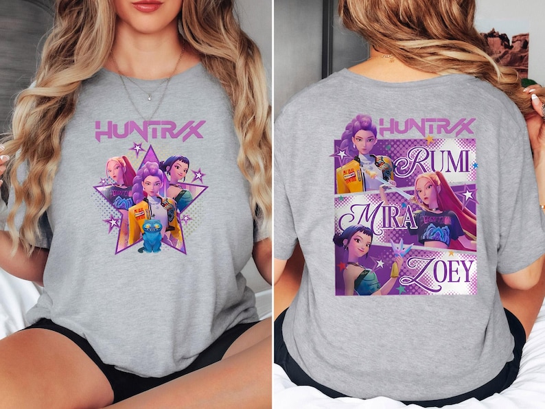 May include: A heather gray t-shirt featuring a graphic of a star with animated characters and the text "HUNTRAX". The back of the shirt displays the names Rumi, Mira, and Zoey with character illustrations.