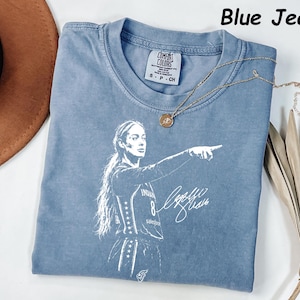 May include: A blue jean colored t-shirt featuring a white graphic print of a basketball player pointing, with a signature and the text Blue Jean. The shirt has a Comfort Colors label at the neck and is paired with a gold pendant necklace.