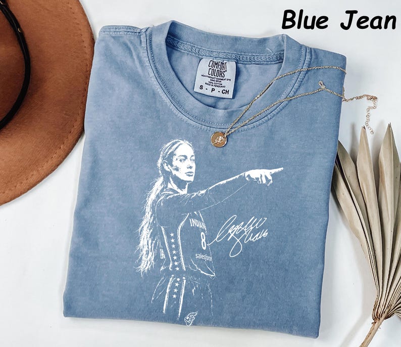 May include: A blue jean colored t-shirt featuring a white graphic print of a basketball player pointing, with a signature and the text Blue Jean. The shirt has a Comfort Colors label at the neck and is paired with a gold pendant necklace.