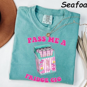 May include: Seafoam green t-shirt with the text "PASS ME A FRIDGE CIG" in pink. The graphic is a box of pink and white cans. A gold necklace is draped on the shirt.